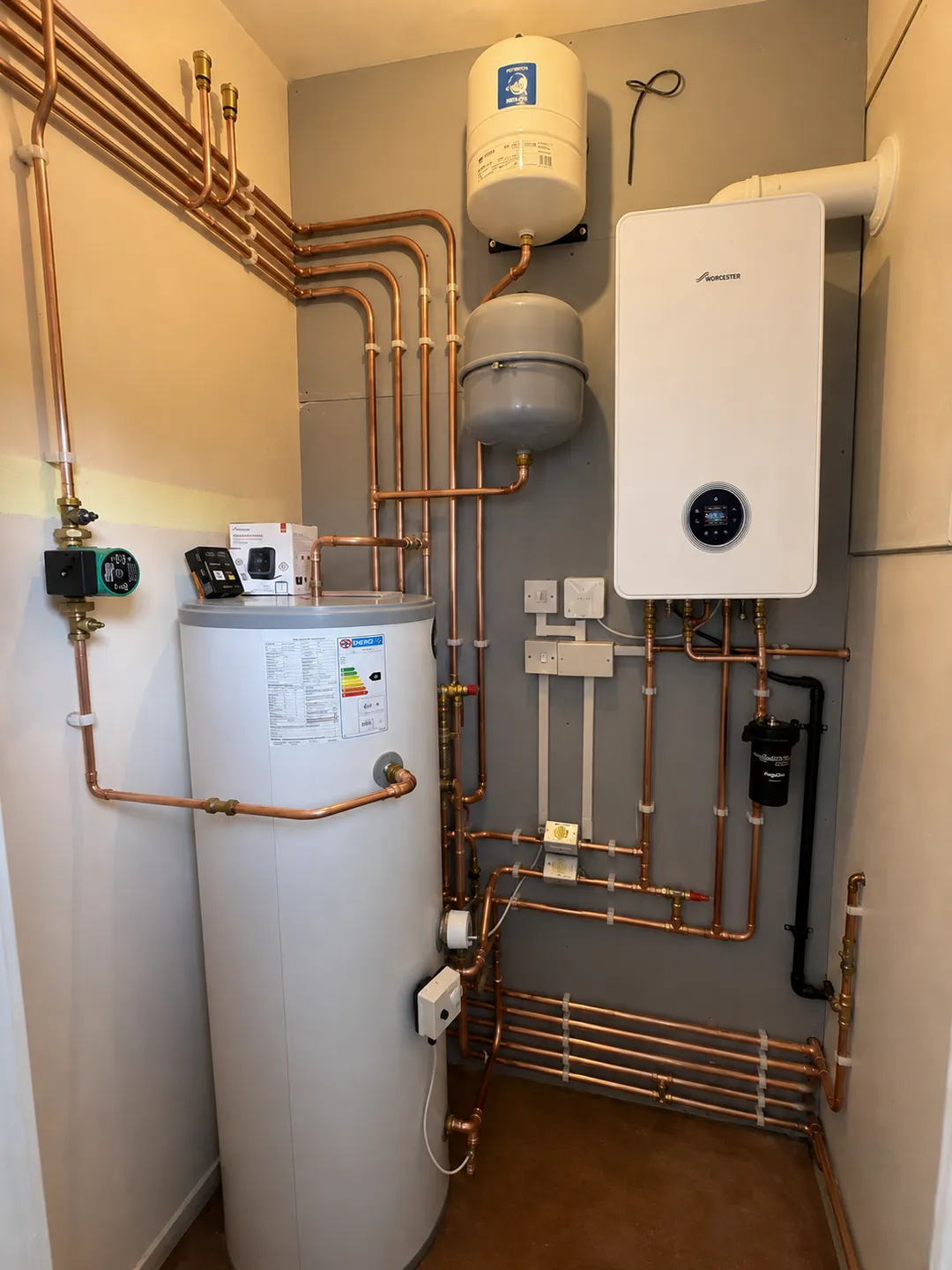 A plant room fitted by On Heating: a wall mounted Worcester system boiler, an unvented hot water cylinder, two expansion vessels, a pump and a magnetic filter, connected in copper pipework.